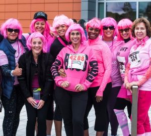 A group of women wearing pink wigs and pink shirts pose together at the conclusion of a 5K race in support of those with breast cancer.