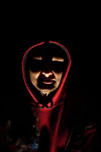 A person wearing a red hood whose face is lit from underneath by a light making it hard to see their features.