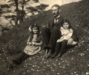 Informal photograph of JMcC sitting on a flowery bank with his two youngest daughters, Joan (left, b 1911) and Peggy (right, 1913).