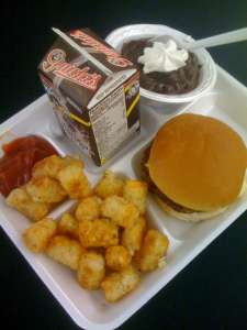 Classic school lunch containing a hamburger, tater tots with ketchup, chocolate pudding, and chocolate milk.