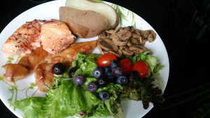 Sample healthy meal for an athlete. It contains fish and poultry, starchy potato, mushrooms, and a garden salad with tomatoes and blueberries.