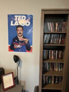 Shelves with DVDs and a poster of Ted Lasso holding a cup of tea.