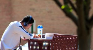 A male student wearing headphones sits at an outdoor table writing. On the table are a book and a water bottle.